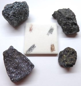 Four minerals around a white streak plate. Each mineral has been scratched on the plate leaving a colored powder.