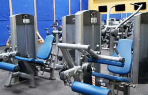 Exercise training room with stacked weight machines in grey color with blue pads. The leg extension and leg curl are in focus, with other machines in the background.