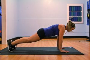 A woman in black shorts and a purple shirt performs a plank exercise on a black mat on a wooden floor.